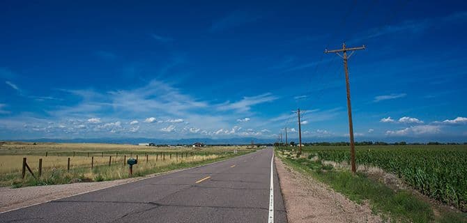 A rural road stretches into the distance between fields under a clear blue sky with scattered clouds. Power lines run alongside the road.