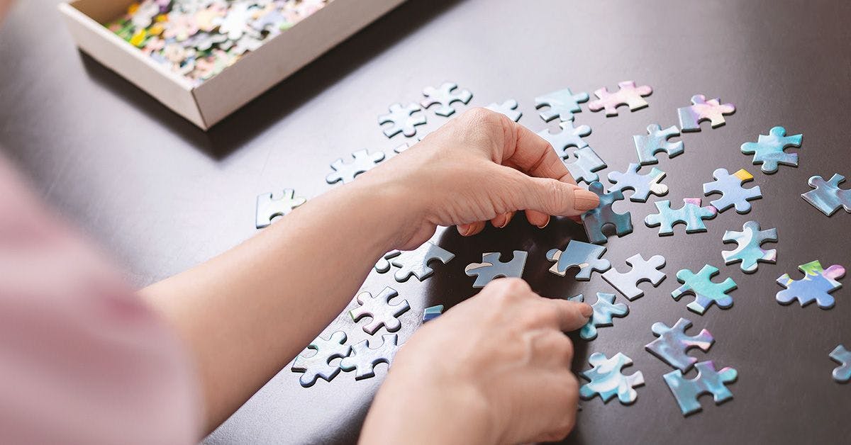 Two hands assembling a jigsaw puzzle on a table, with scattered pieces and a puzzle box nearby.