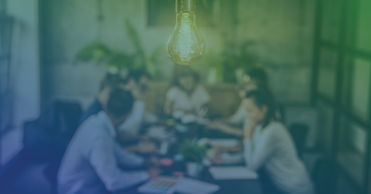 A team of professionals engaged in a meeting around a table with a glowing lightbulb hanging overhead, symbolizing ideas and innovation.