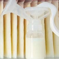 A breast pump with a bottle containing milk is placed in front of several stored bags of breast milk.