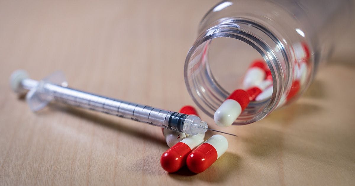 A syringe lies beside a tipped-over glass bottle with red and white capsules spilling out on a wooden surface.