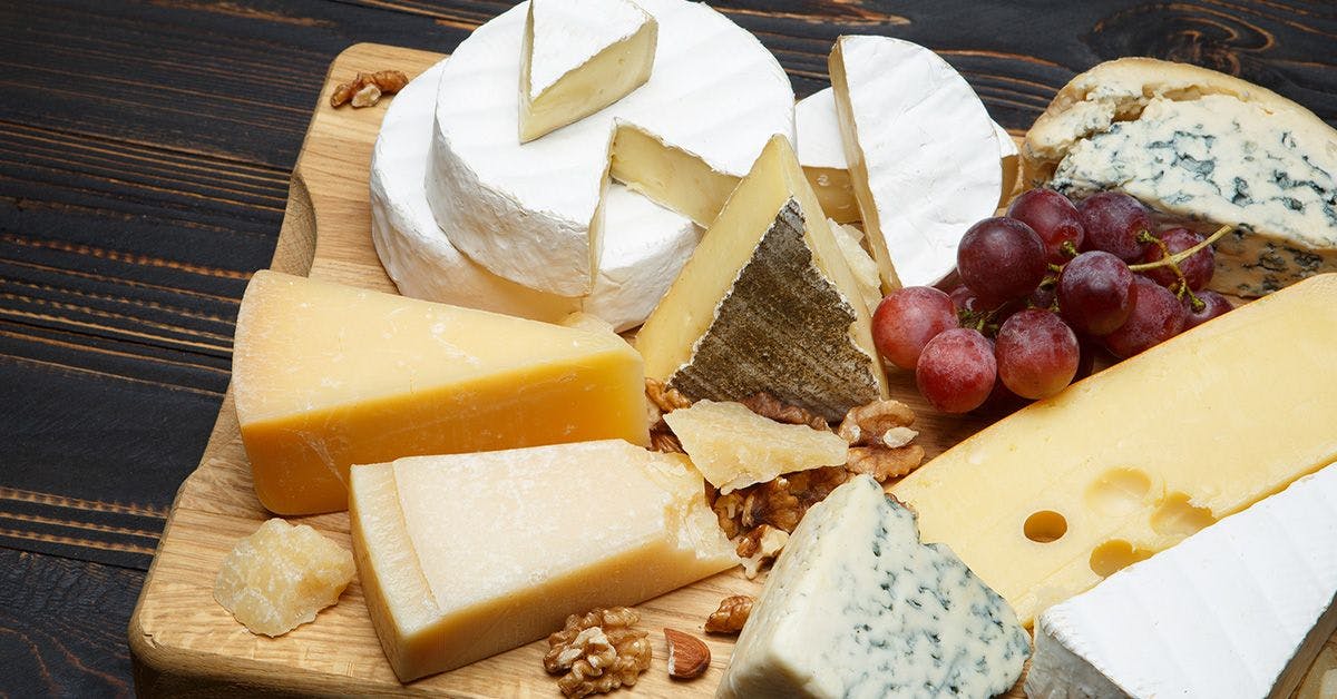 A wooden board with assorted cheeses including brie, blue cheese, cheddar, and Swiss, accompanied by red grapes and walnuts.
