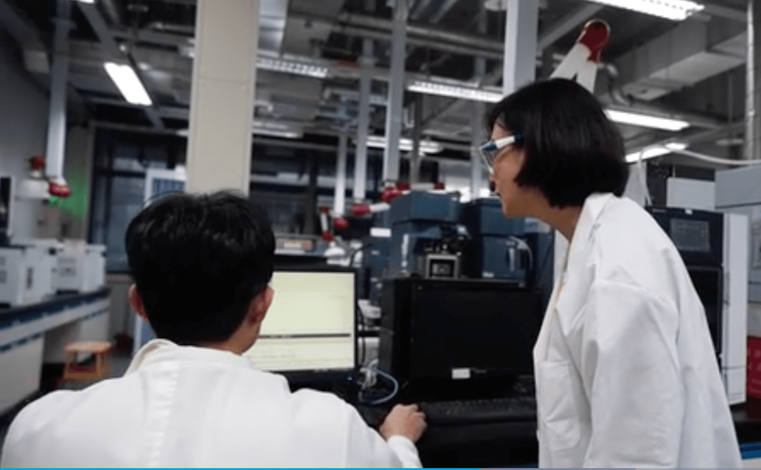 Two individuals in lab coats are working at a computer in a laboratory. The person on the left is seated and typing on the keyboard, while the person on the right is standing and observing the screen.