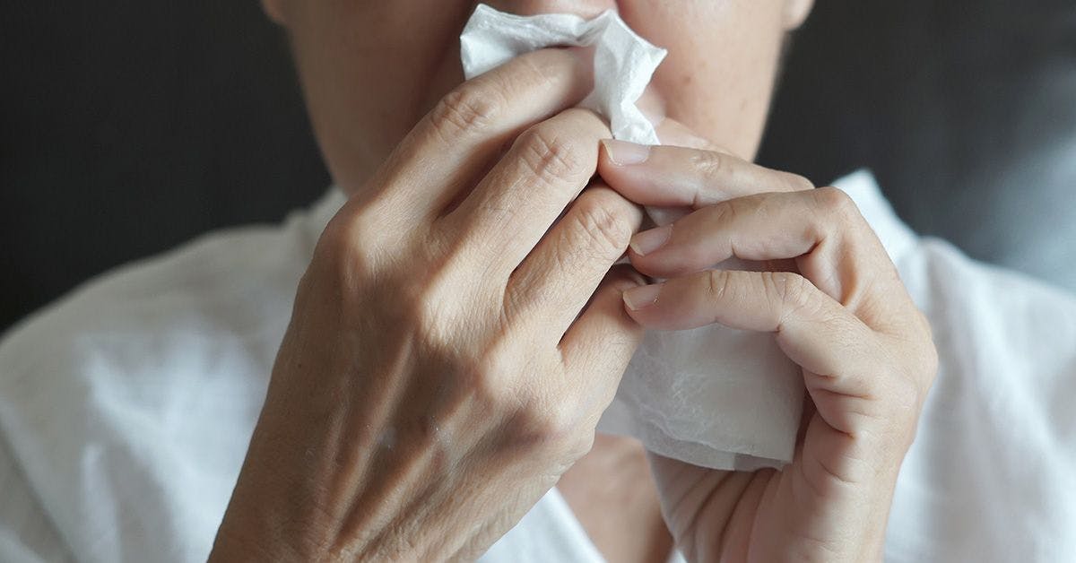 A person, partially visible, holds a tissue to their nose, appearing to sneeze or blow their nose.