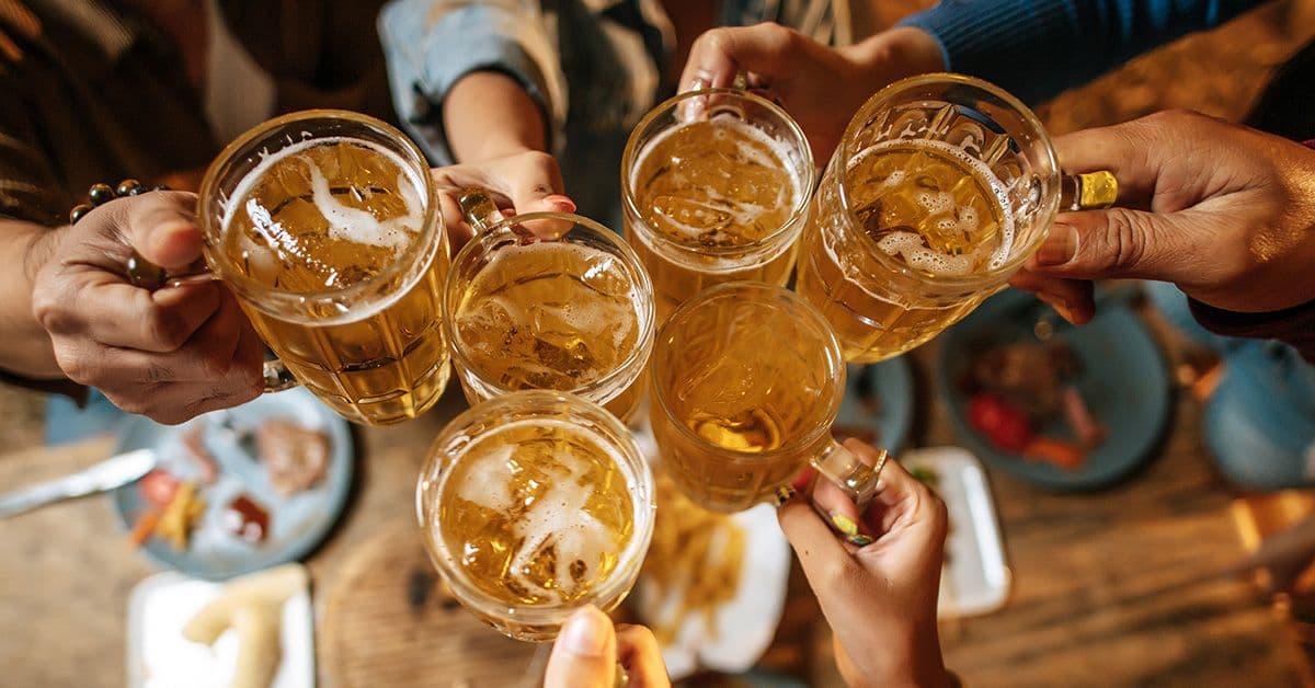 Six people clinking glasses of beer over a wooden table, viewed from above.