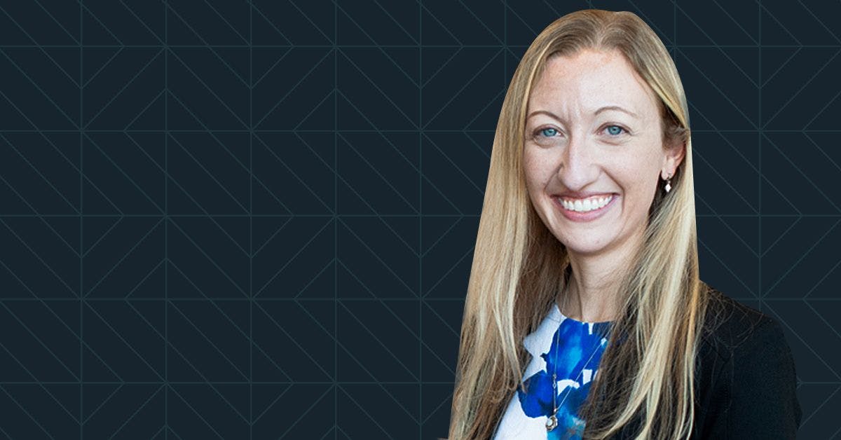 A woman with long blonde hair, wearing a black blazer and a blue and white top, is smiling against a dark geometric patterned background.