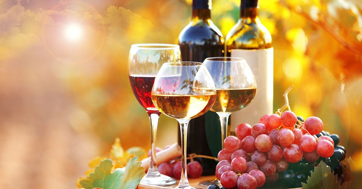 Three wine glasses filled with red and white wine, two wine bottles, and a bunch of grapes on a table outdoors.