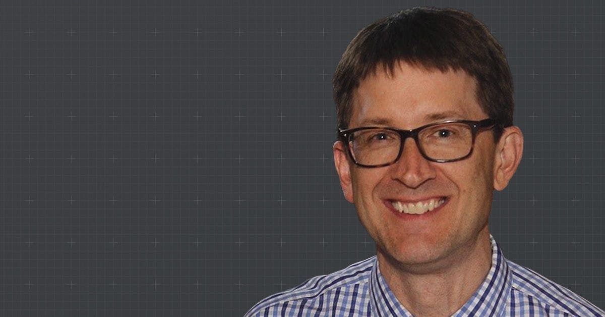 A smiling man with glasses and short hair wearing a checkered shirt against a dark, textured background.