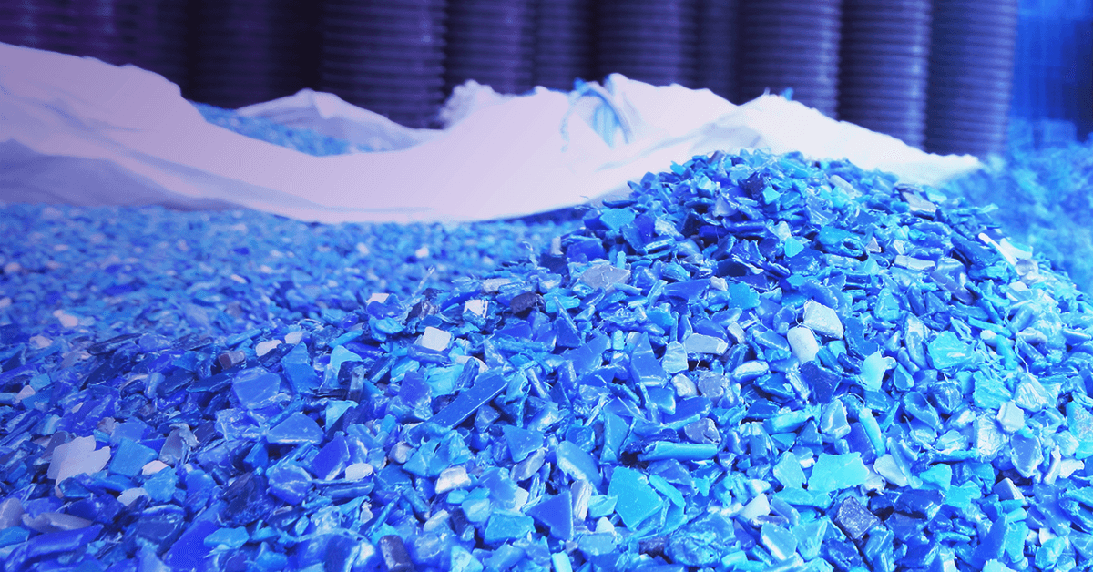 A pile of blue plastic fragments sits in front of stacked containers in a warehouse setting.