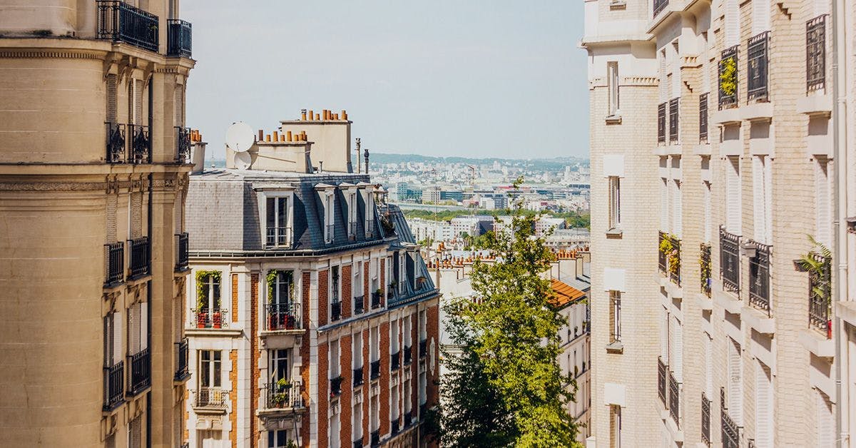 Parisian cityscape viewed between buildings, showcasing traditional French architecture with mansard roofs, balconies, and greenery in the distance under a clear sky.