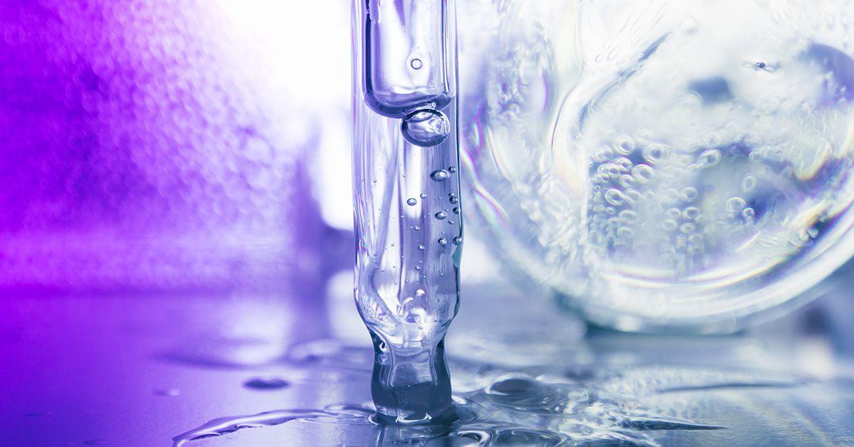 Close-up of a vertical test tube with clear liquid and bubbles inside, surrounded by a wet surface and a blurred laboratory background with a purple hue.