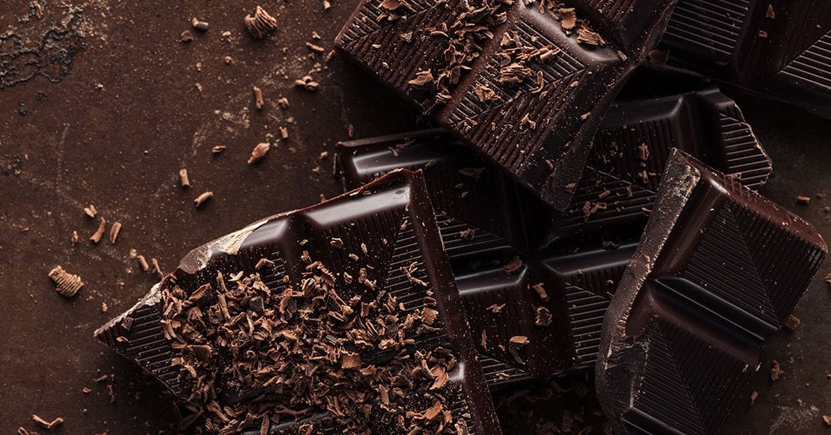 pieces of a dark chocolate bar and chocolate shavings scattered on a dark brown background.