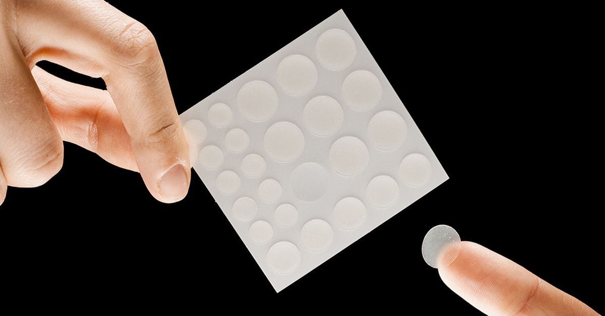 A sheet of hydrogel pimple patches against a black background; one hand holding the sheet and one hand removing a patch.