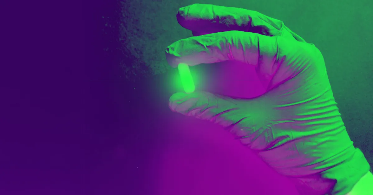 Scientific-style image showing a gloved hand holding a bright green capsule between fingers, with dramatic purple and green lighting effects.