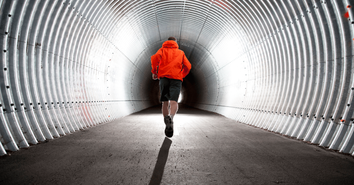 A person wearing an orange jacket moves through a ribbed metal tunnel, with light visible at the far end.