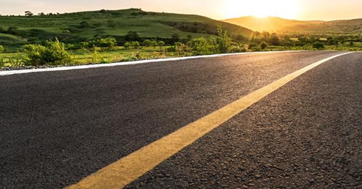 A Low-angle view of an empty asphalt road with a yellow centerline stretching into green hills, under a clear blue sky with the sun rising on the horizon.