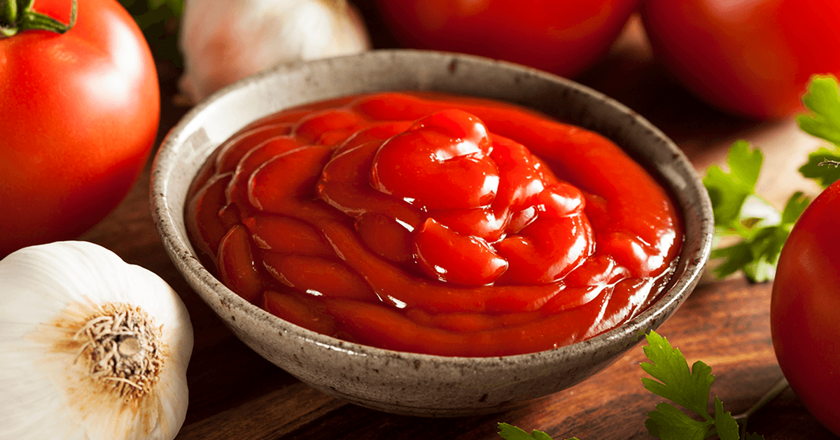 Close-up of a bowl filled with bright red ketchup, surrounded by whole tomatoes, garlic bulbs, and parsley on a wooden surface.