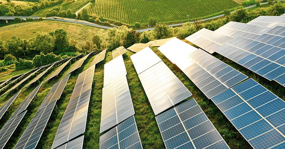 An aerial view of long rows of solar panels installed on a grassy hillside, surrounded by trees, farmland, and winding roads.