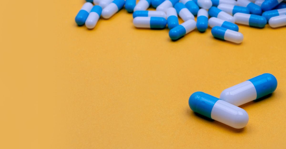 Several blue and white capsules are scattered on a yellow surface, with two capsules prominently placed in the foreground.