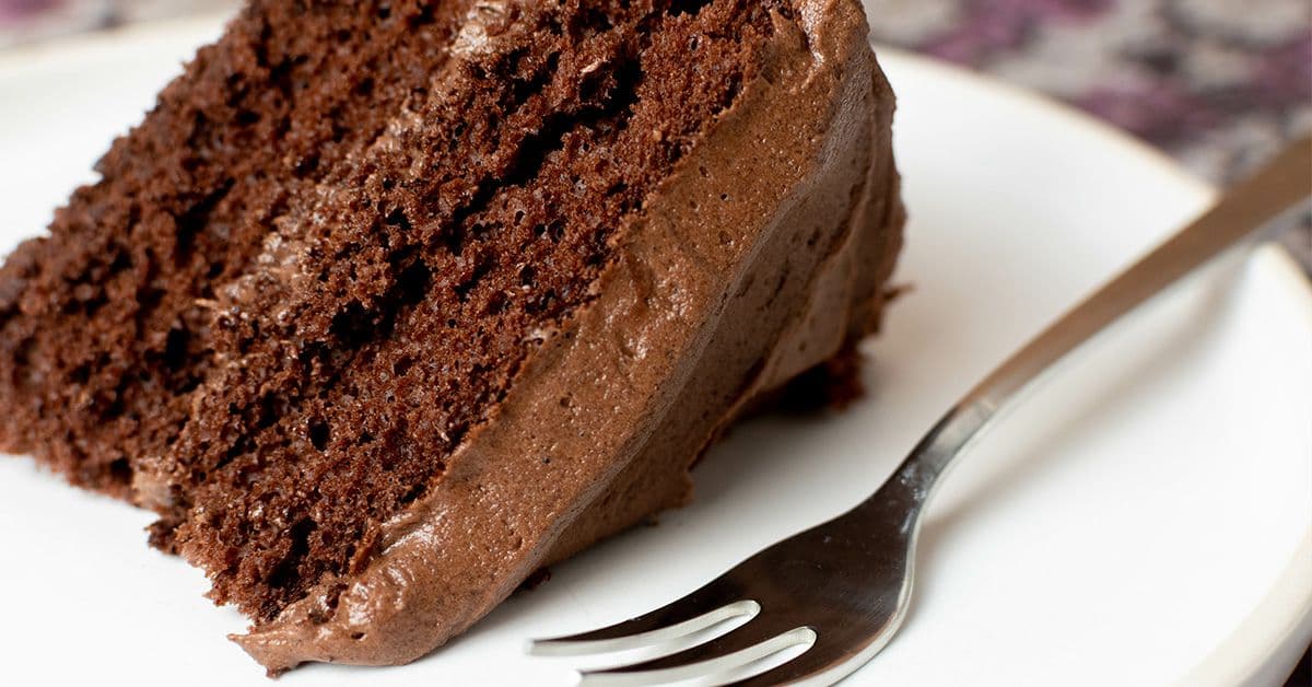A slice of chocolate cake with creamy frosting, placed on a white plate beside a fork.
