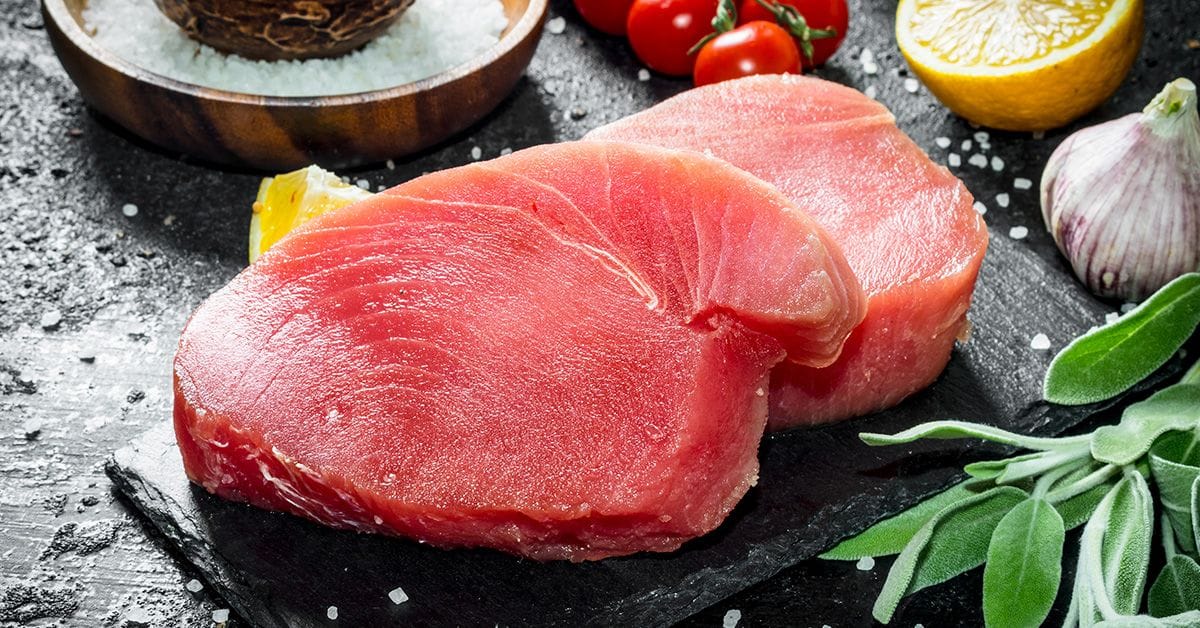 Two fresh, pink tuna steaks on a black slate with herbs, garlic, cherry tomatoes, lemon, and a wooden bowl of salt in the background.