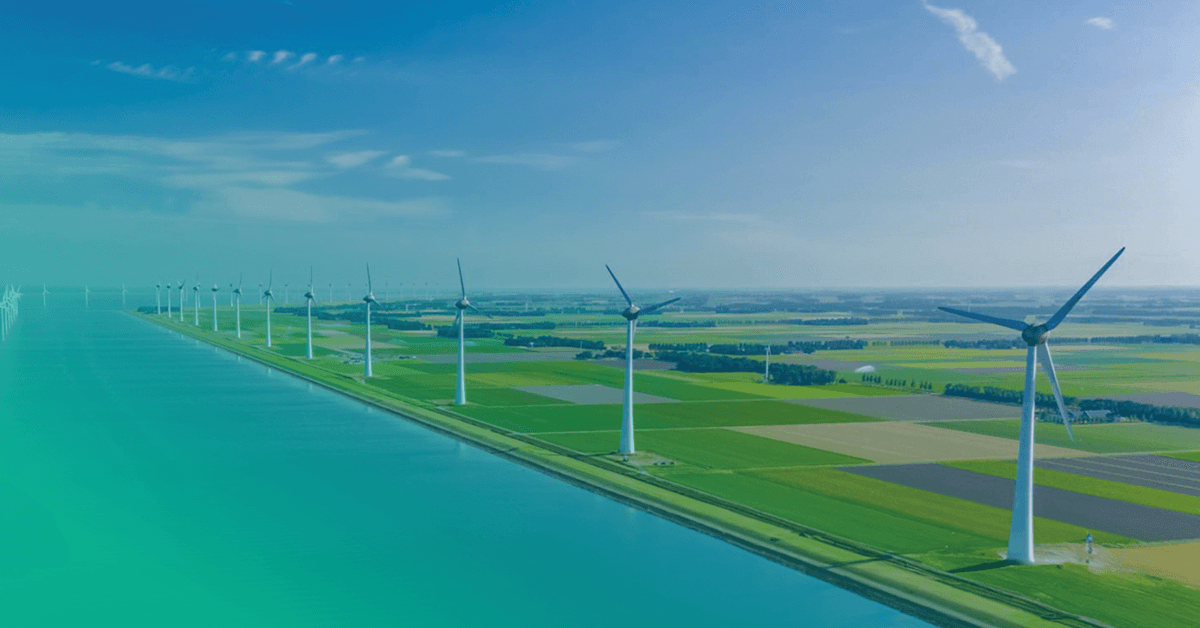 A row of wind turbines positioned along a coastline, with green fields and a body of water visible under a clear blue sky.