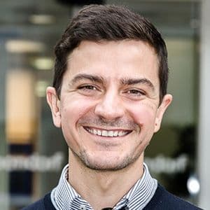 A smiling man with short dark hair, wearing a checkered shirt and sweater, standing outdoors in front of a glass building.