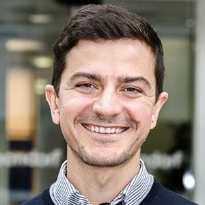 A smiling man with short dark hair, wearing a checkered shirt and sweater, standing outdoors in front of a glass building.