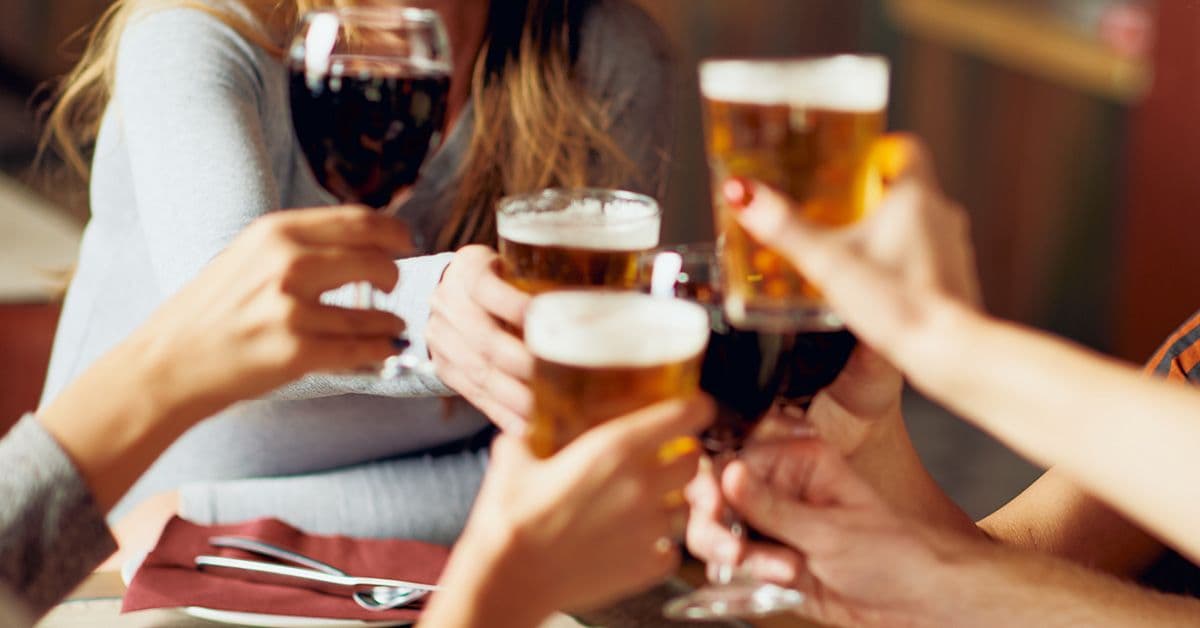 Group of people raising glasses of beer and wine in a toast at a table, with a napkin and cutlery visible.