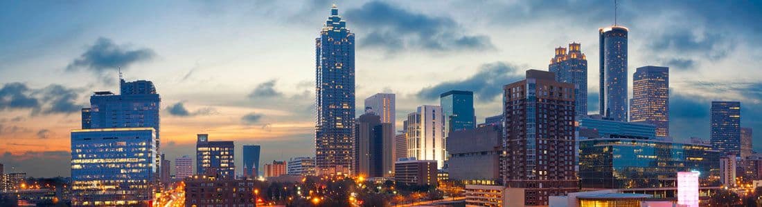 Skyline of a city at dusk with illuminated high-rise buildings and a cloudy sky.