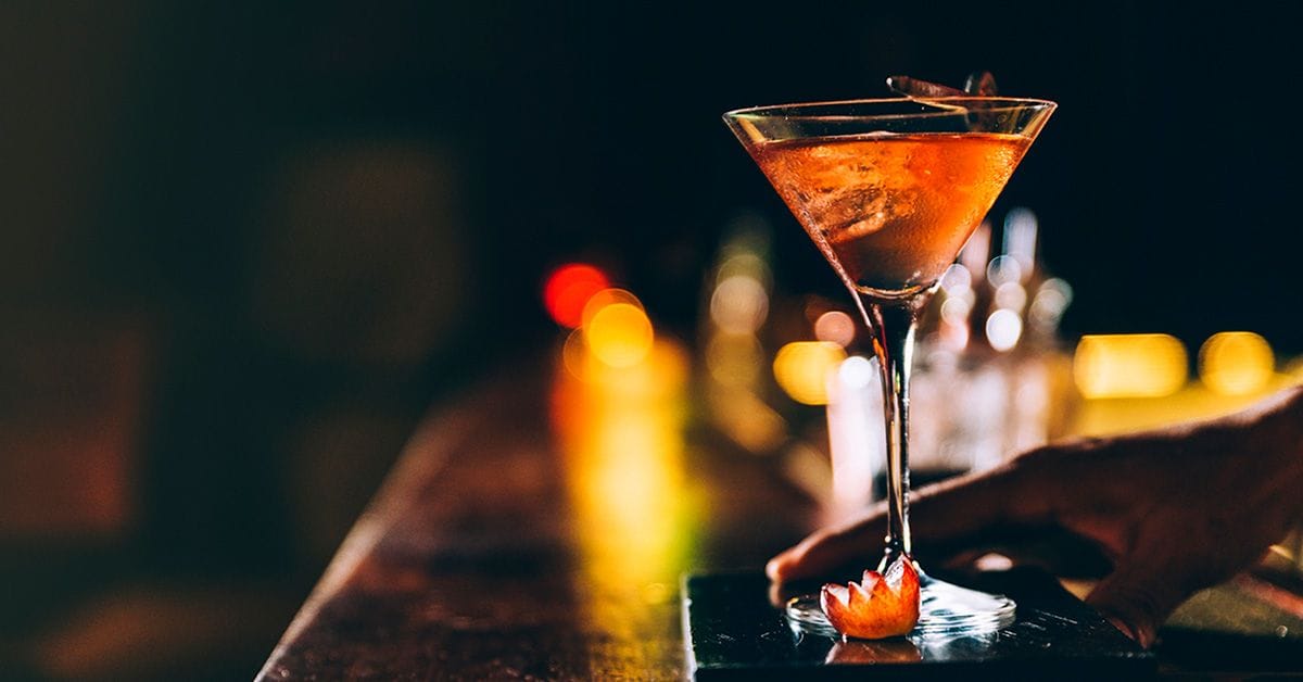 An image of a bartender's hand serving an elegant mixed cocktail across a bar countertop, set against a dark, blurred background.