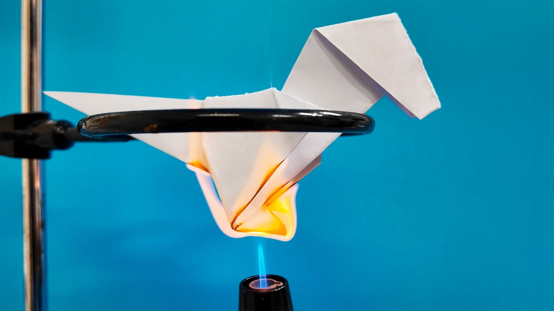 A laboratory setup showing a folded white origami paper t-rex held in a metal clamp while a burner flame heats the underside, suggesting thermal testing.