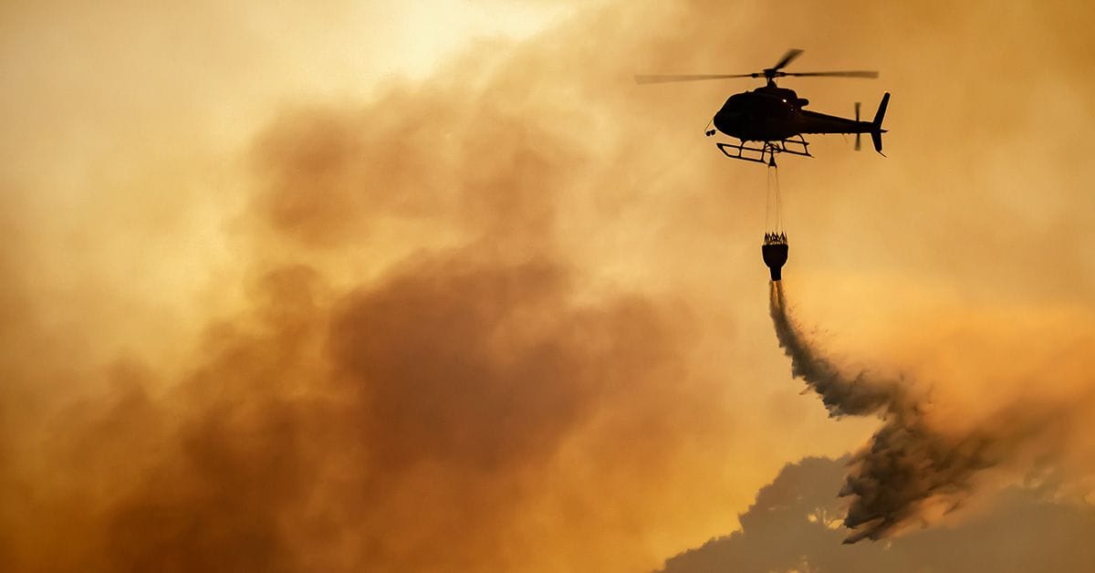 A helicopter releases water over a smoky wildfire area, with an orange sky in the background.