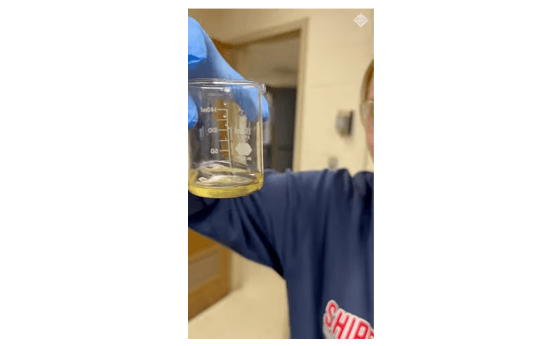 Person in a blue glove holding a beaker with a yellow liquid.