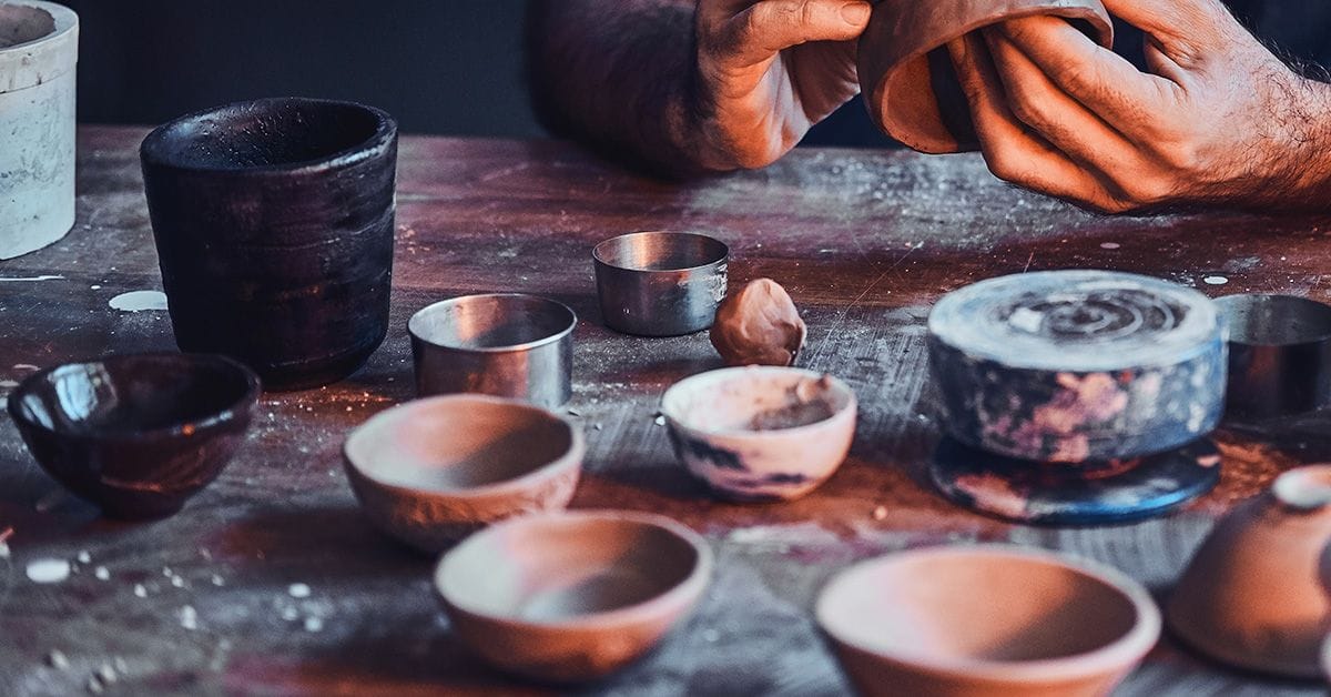 Hands shaping a clay bowl on a table with various ceramic bowls and materials scattered around.