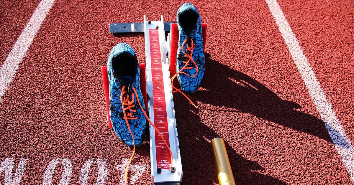 A pair of blue track spikes with orange laces are placed on a red running track next to a white starting block and a relay baton.