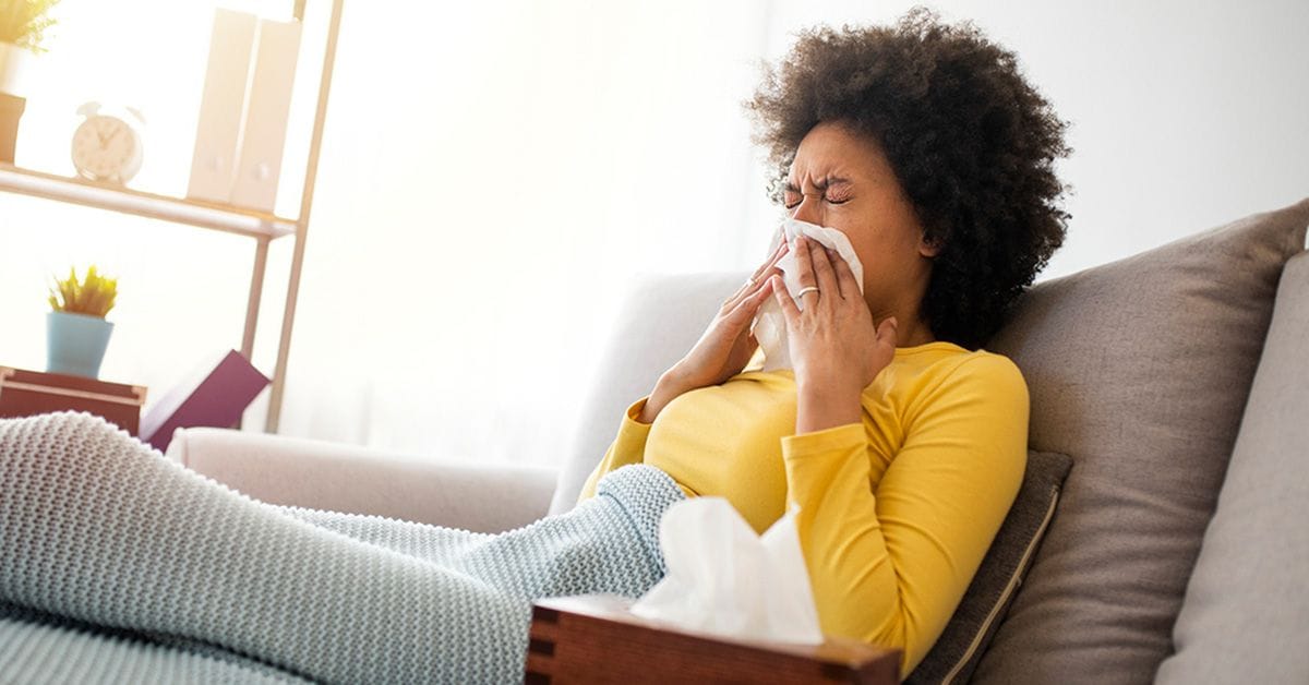 A woman with the flu reclining on a couch with a blanket and blowing her nose, with a box of tissues nearby.
