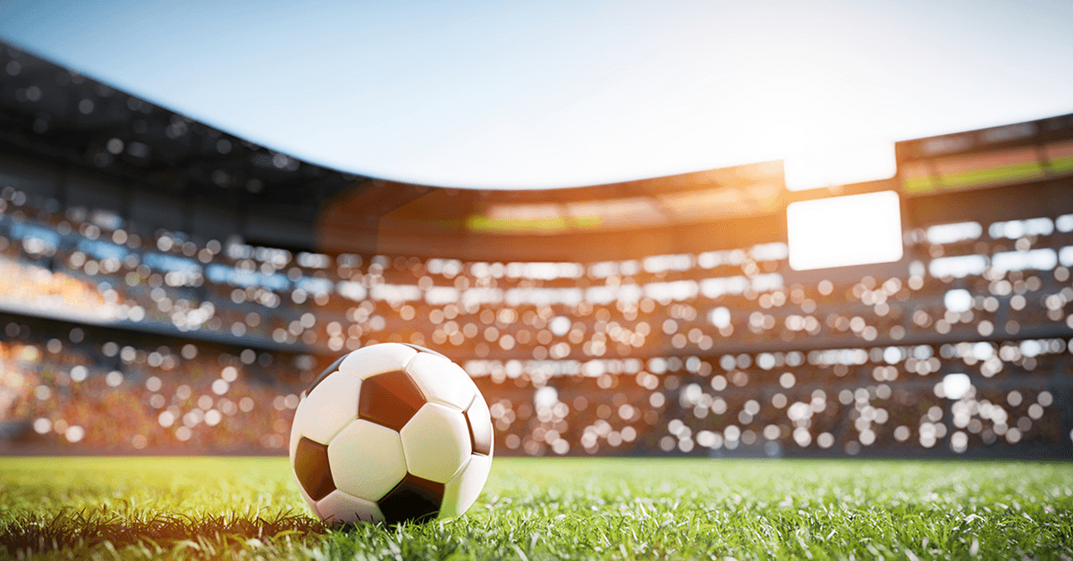 Close-up of a soccer ball on a grassy field, with stadium lights and spectators softly blurred in the background during a match.