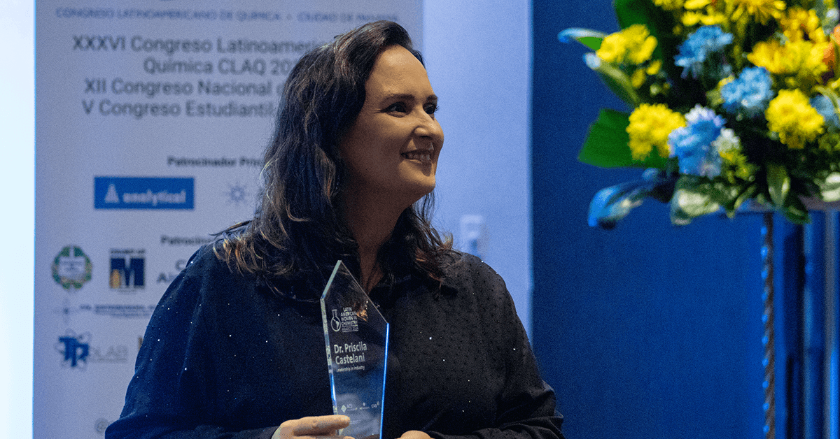 A person holding a glass award stands smiling in front of a conference backdrop and a flower arrangement.