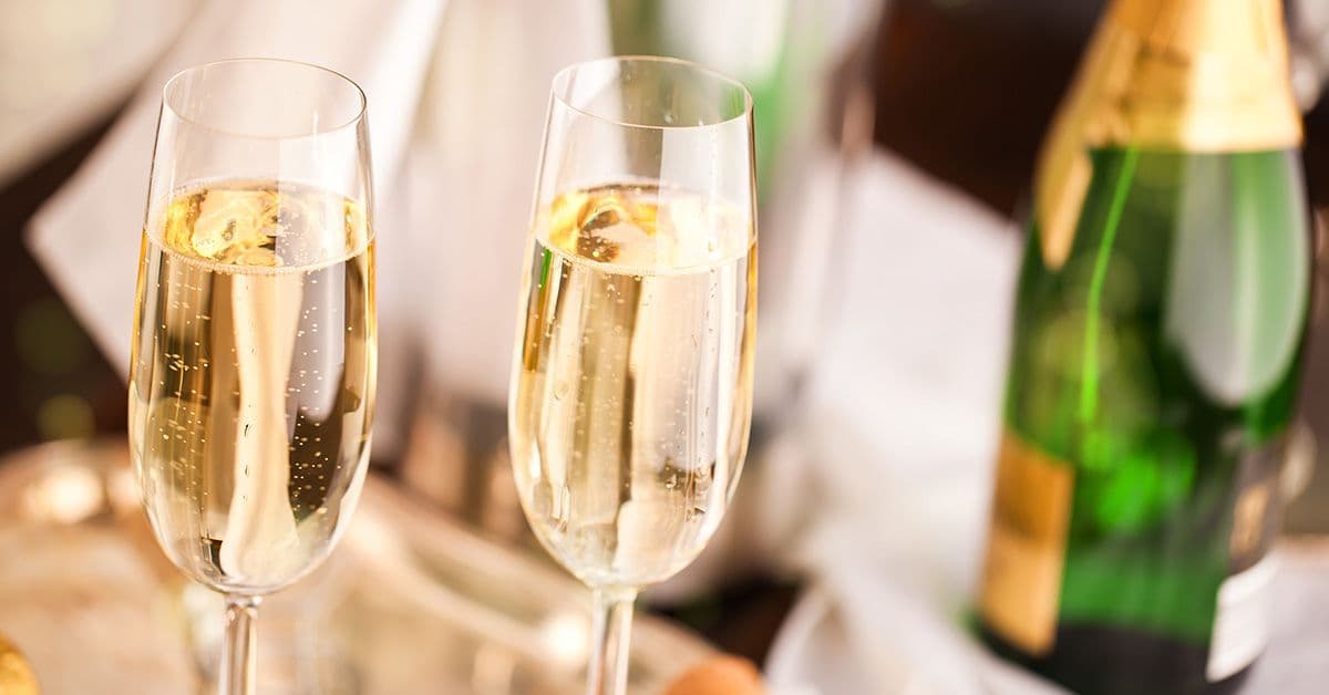 Two champagne glasses filled with bubbling liquid are in focus, with blurred bottles in the background.