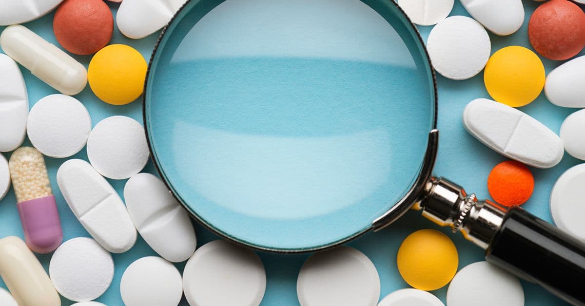 A magnifying glass over a variety of colorful pills and capsules scattered on a blue background.