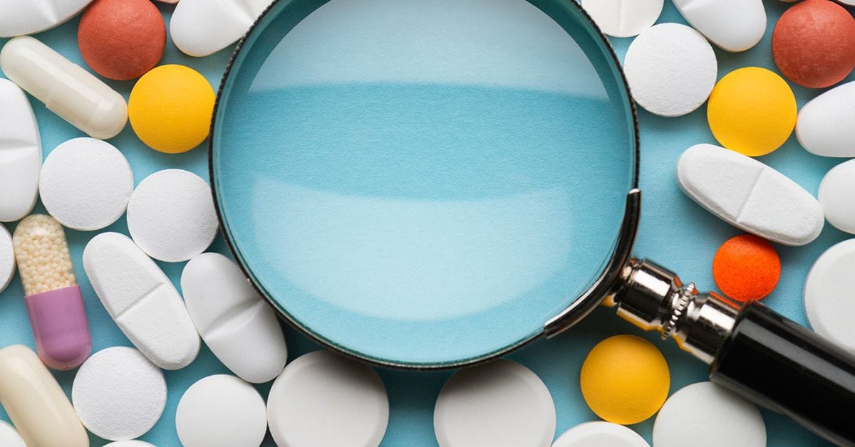 A magnifying glass over a variety of colorful pills and capsules scattered on a blue background.