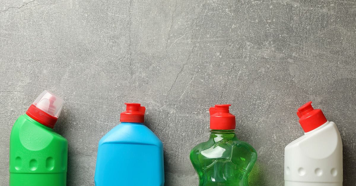 Four colorful plastic detergent bottles with red caps are aligned against a gray, textured background. The bottles are green, blue, another green, and white.