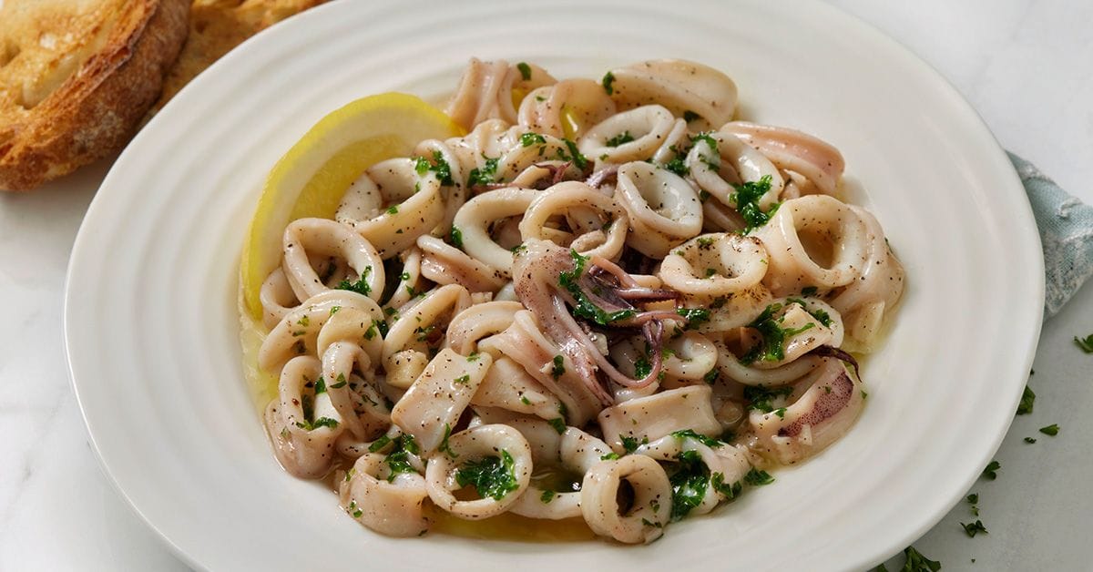 A plate of cooked calamari garnished with parsley and lemon, served with slices of toasted bread in the background.