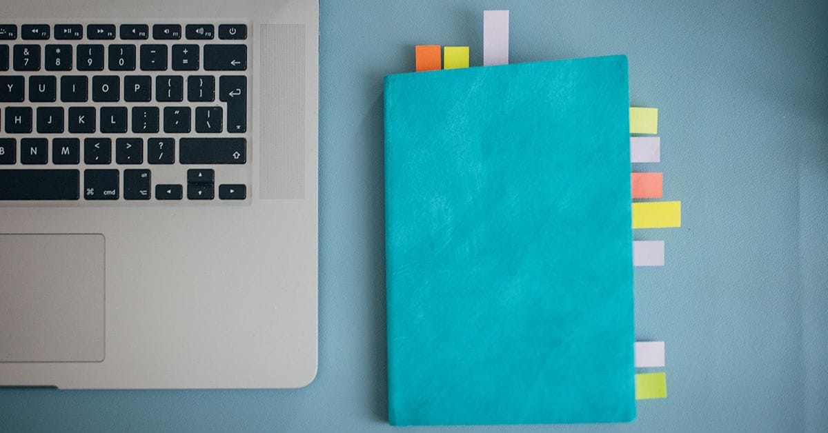 A turquoise notebook with colorful tabs sits beside a MacBook keyboard on a light blue surface.