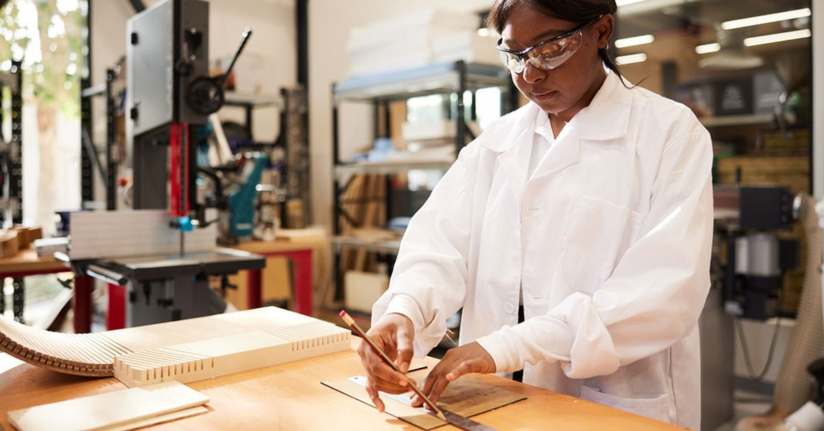 In a white lab coat and protective eyewear, a woman draws precise lines on particleboard.