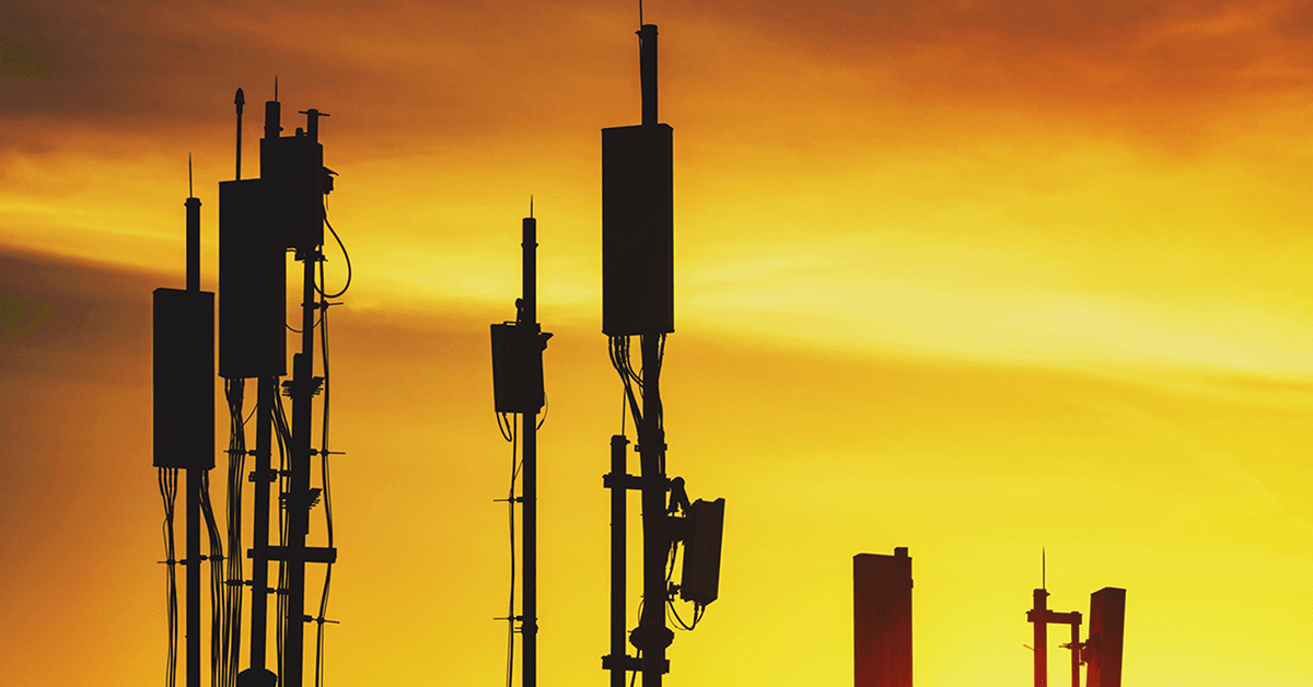 Silhouettes of telecommunications antennas and cell tower equipment against a vivid orange and yellow sunset.