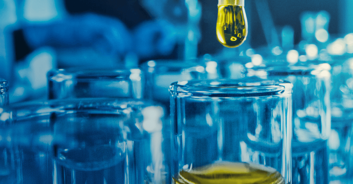 A yellow liquid dropper above a glass container, surrounded by laboratory glassware in a blue-tinted setting.