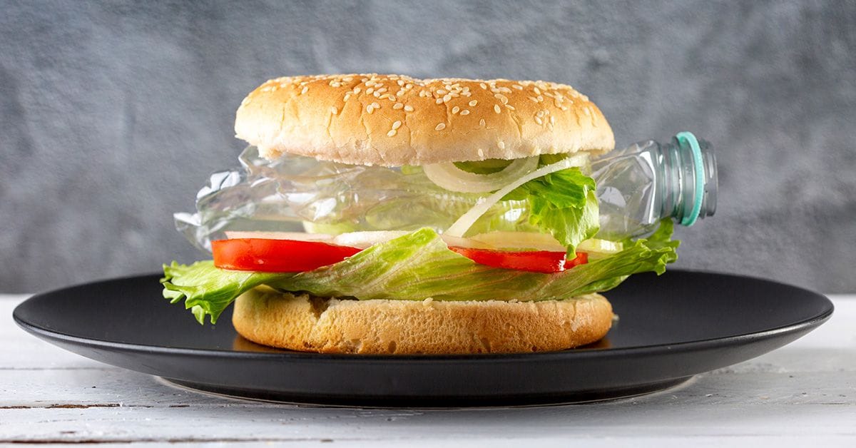 A hamburger made with a sesame bun, leafy lettuce, tomato, and onion, featuring a transparent plastic bottle as the filling.