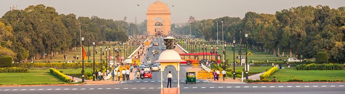A wide road leads to India Gate in New Delhi, lined with trees and people, with vehicles and buses on the road.